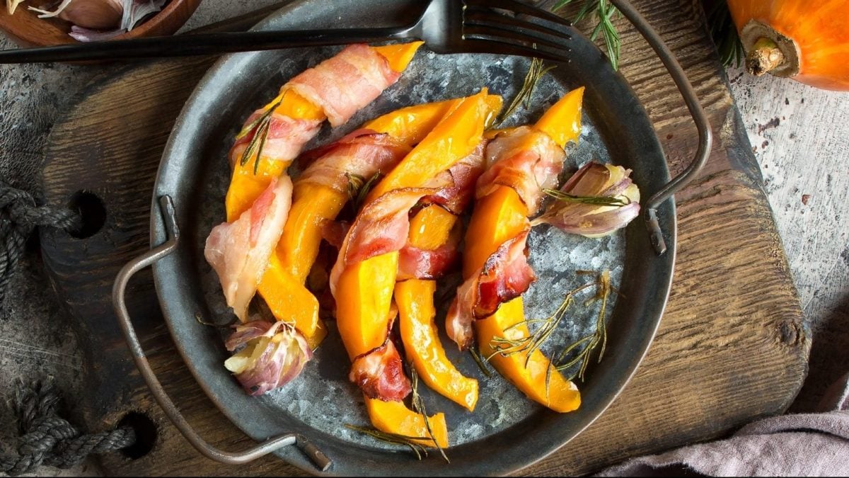 Draufsicht: Eine gusseiserne Pfanne. In der liegen mit Speck bardierte Kürbisspalten.