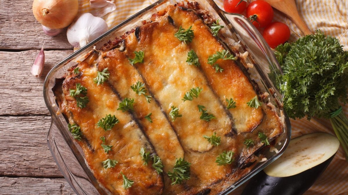 Original griechische Moussaka ohne Kartoffeln in Glasform auf Holz. Im Bild Tomaten, Petersilie und weitere Zutaten. Draufsicht.