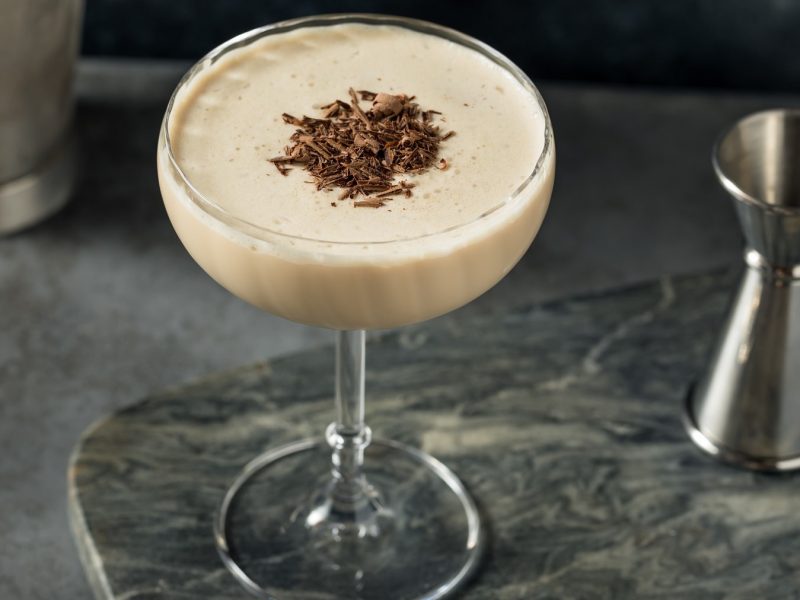 Ein Cocktailglas mit dem Mudslide-Cocktail auf grauem Untergrund. Daneben ein silberner Shaker und ein Jigger.