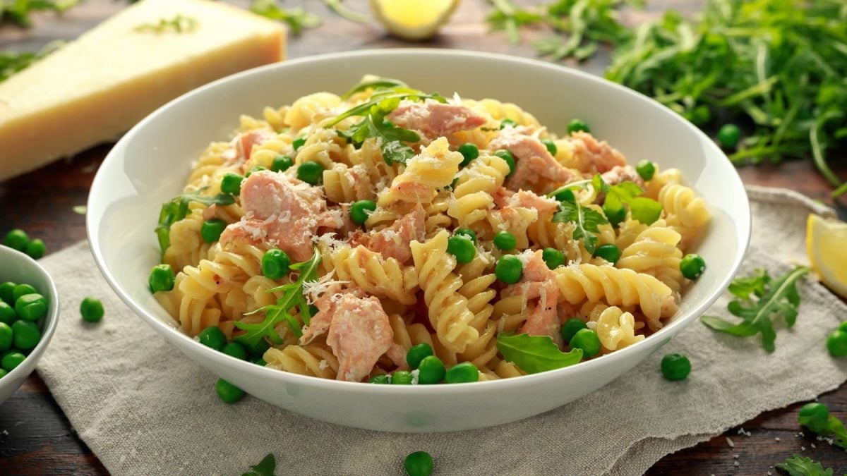 Ein weißer, tiefer Teller mit dem Nudelsalat mit Thunfisch und Erbsen auf heller Serviette, aber dunklem Holztisch. Im Hintergrund sind einige Salatzutaten unscharf zu erkennen. Alles seitlich fotografiert.