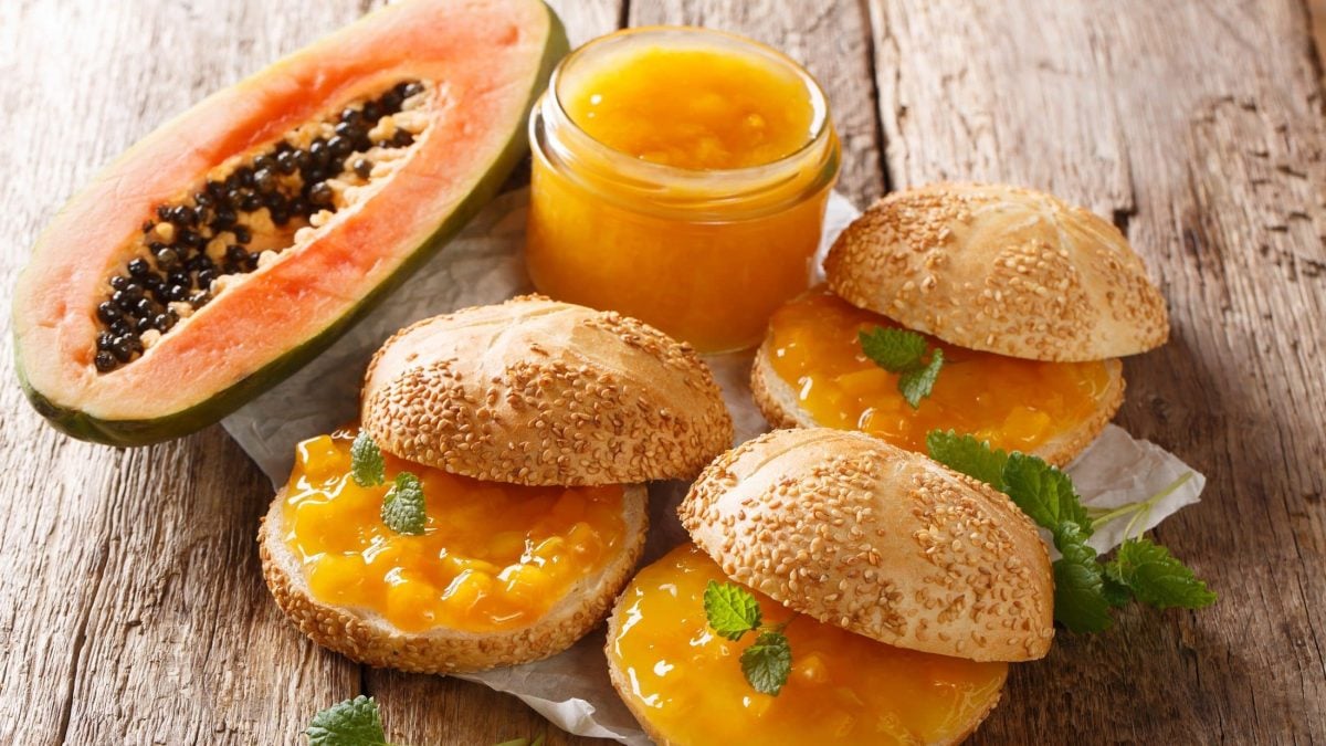 Draufsicht schräg: Ein Glas Papaya-Aufstrich, drei Sesambrötchen mit Papaya-Aufstrich und eine halbe, aufgeschnittene Papaya.
