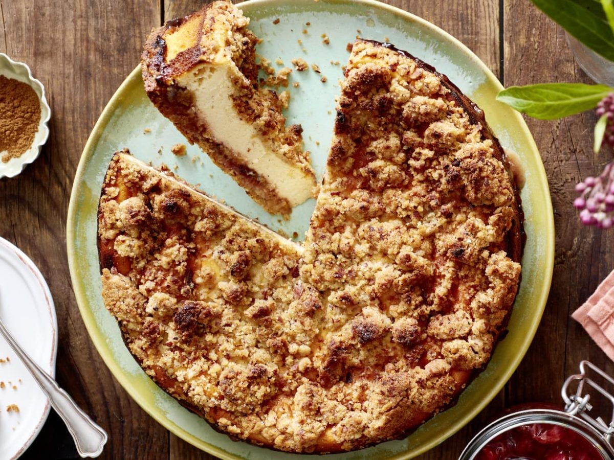 Ein Pflaumen-Käsekuchen auf einem grünen Servierteller auf einem hölzernen Hintergrund. Links davon ein Teller mit einem Stück Kuchen und einer Dessertgabel, darüber eine kleine Schüssel mit Zimt. Rechts Blumen, ein Geschirrtuch und ein Bügelverschlussglas mit Kompott.