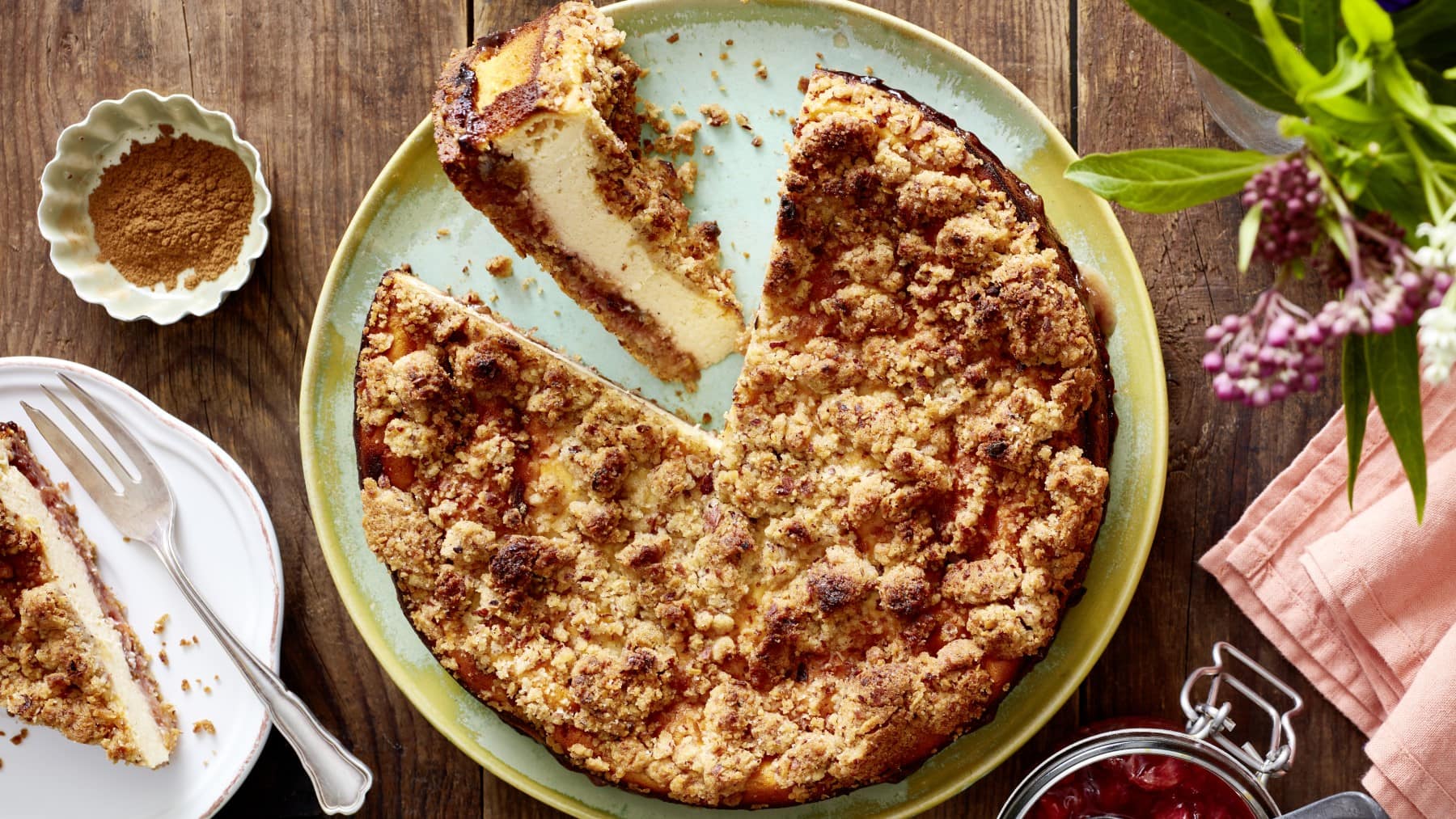 Ein Pflaumen-Käsekuchen auf einem grünen Servierteller auf einem hölzernen Hintergrund. Links davon ein Teller mit einem Stück Kuchen und einer Dessertgabel, darüber eine kleine Schüssel mit Zimt. Rechts Blumen, ein Geschirrtuch und ein Bügelverschlussglas mit Kompott.