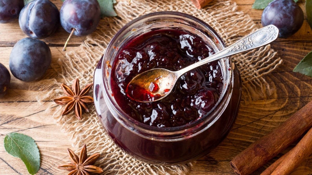 Selbst gemachtes Pflaumenmus im Glas mit kleinem Dessertlöffel auf Leinentuch und Holz. Im Bild Sternanis, Zimt, Blätter und frische Pflaumen. Draufsicht.