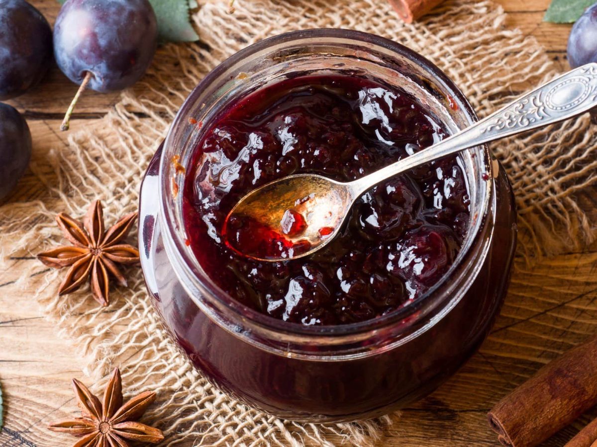 Selbst gemachtes Pflaumenmus im Glas mit kleinem Dessertlöffel auf Leinentuch und Holz. Im Bild Sternanis, Zimt, Blätter und frische Pflaumen. Draufsicht.
