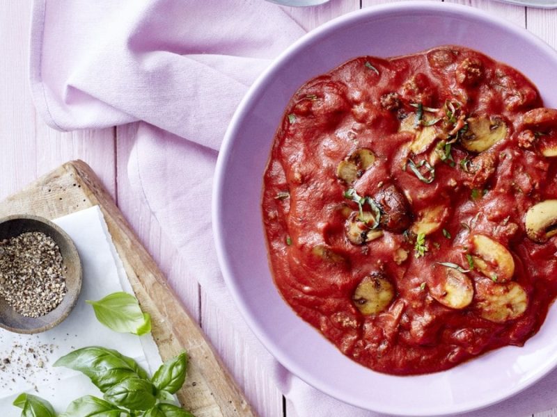 Eine Bolognese mit Pilzen in einem violetten Teller auf einem violetten Geschirrtuch auf einem violetten Hintergrund. Links ein Schneidebrett mit Pfeffer und Basilikum.