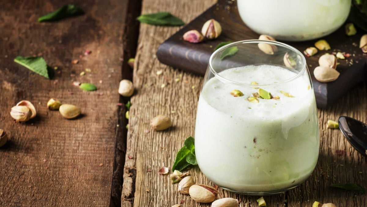 Draufsicht, schräg: ein ganzes und ein halbes Glas mit einem Pistazien-Bananen-Shake auf einem groben Holzboden. Drum herum liegen ganze Pistazien und einige Minzblätter.