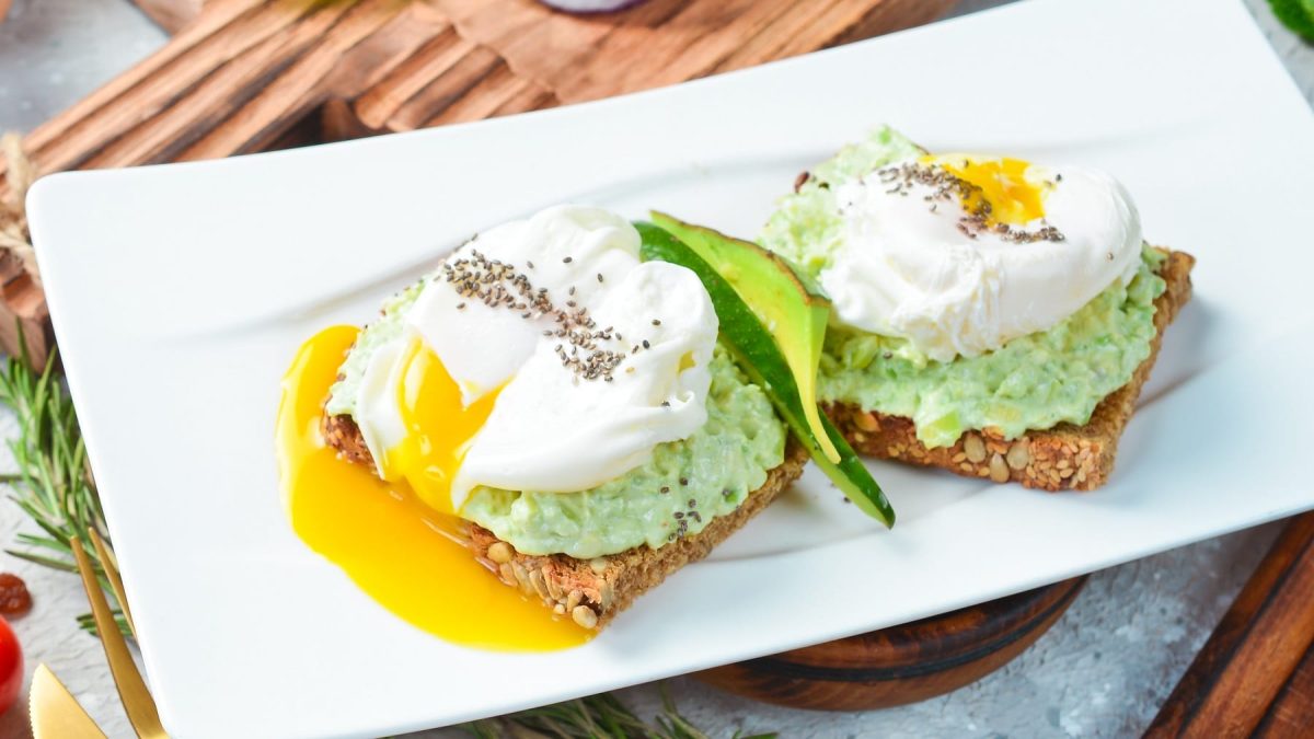Ein weißer, rechteckiger Teller mit dem pochierten Ei auf Avocado-Feta-Creme und dunklem Brot.