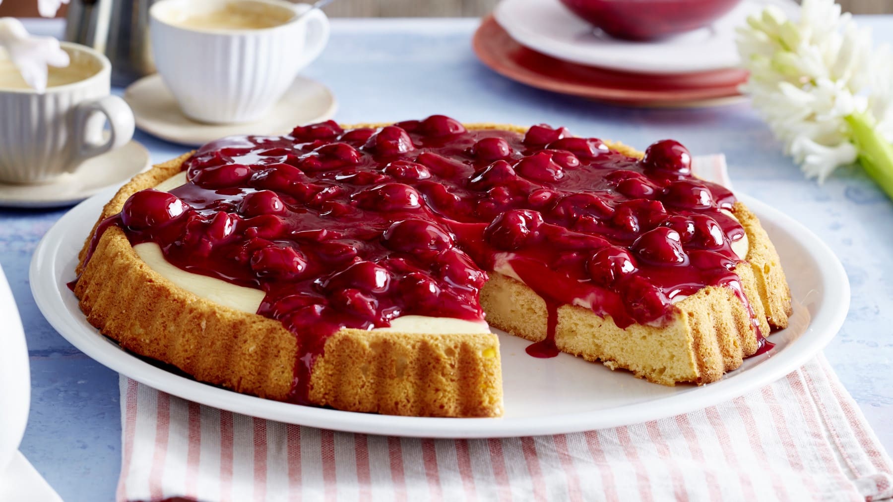 Ein Pudding-Kirschkuchen auf einem weißen Servierteller auf einem weiß-rosa-gestreiftem Geschirrtuch auf einem hellblauem Hintergrund. Links oben Kaffeetassen rechts oben Blumen.