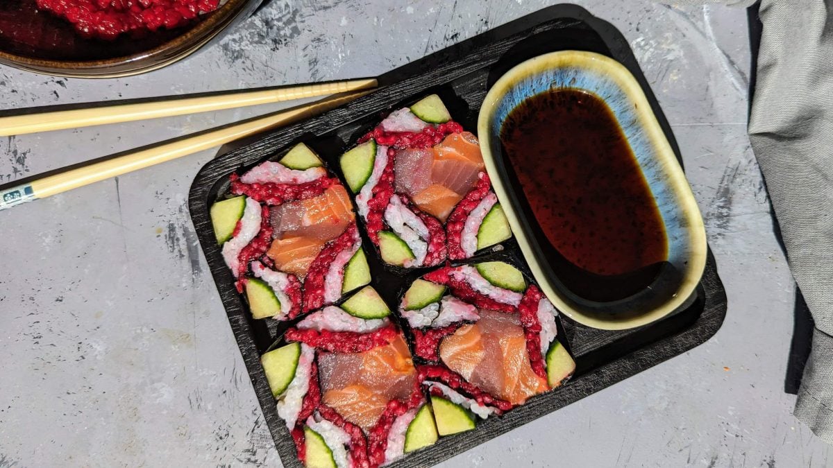 Ein schwarzer viereckiger Teller mit dem Mosaik-Sushi und einer kleinen hellblauen Schale mit Sojasauce von oben fotografiert.