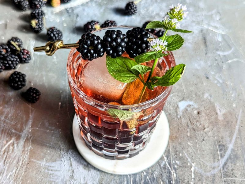 Ein Glas mit dem Brombeer-Smash auf Steinuntergrund. Daneben ein paar Brombeeren und ein kleiner Teil einer Flasche mit Bourbon zu sehen. Alles von oben nach schräg unten fotografiert.