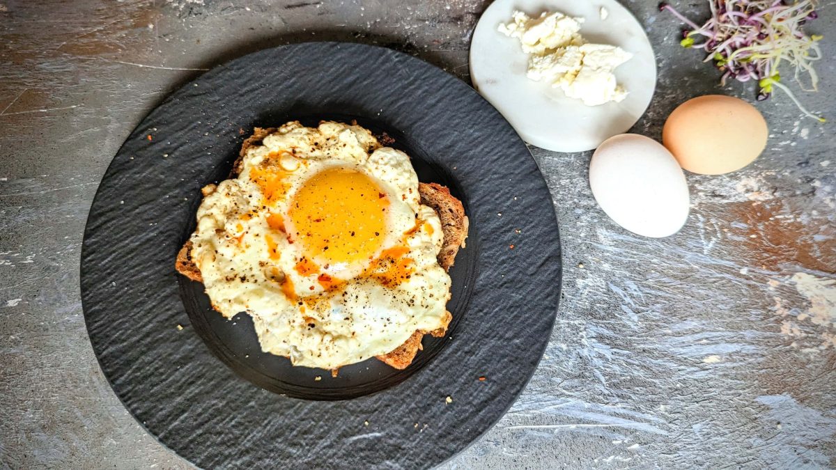 Ein schwarzer Teller mit einer Scheibe Brot und dem Feta-Spiegelei auf Steintisch direkt von oben fotografiert. Rechts oben Feta, Kresse und zwei Eier im Bild.
