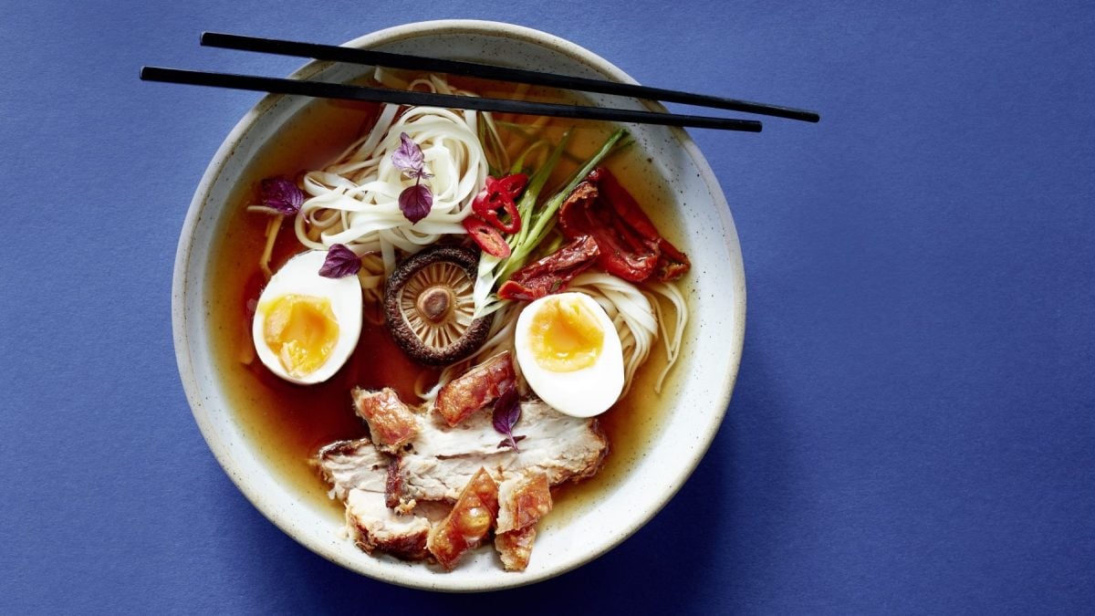 Eine Ramensoup mit Pilz-Dashi und Ei in einer weißen Schüssel vor einem blauen Hintergrund. Auf der Schüssel liegen Stäbchen.