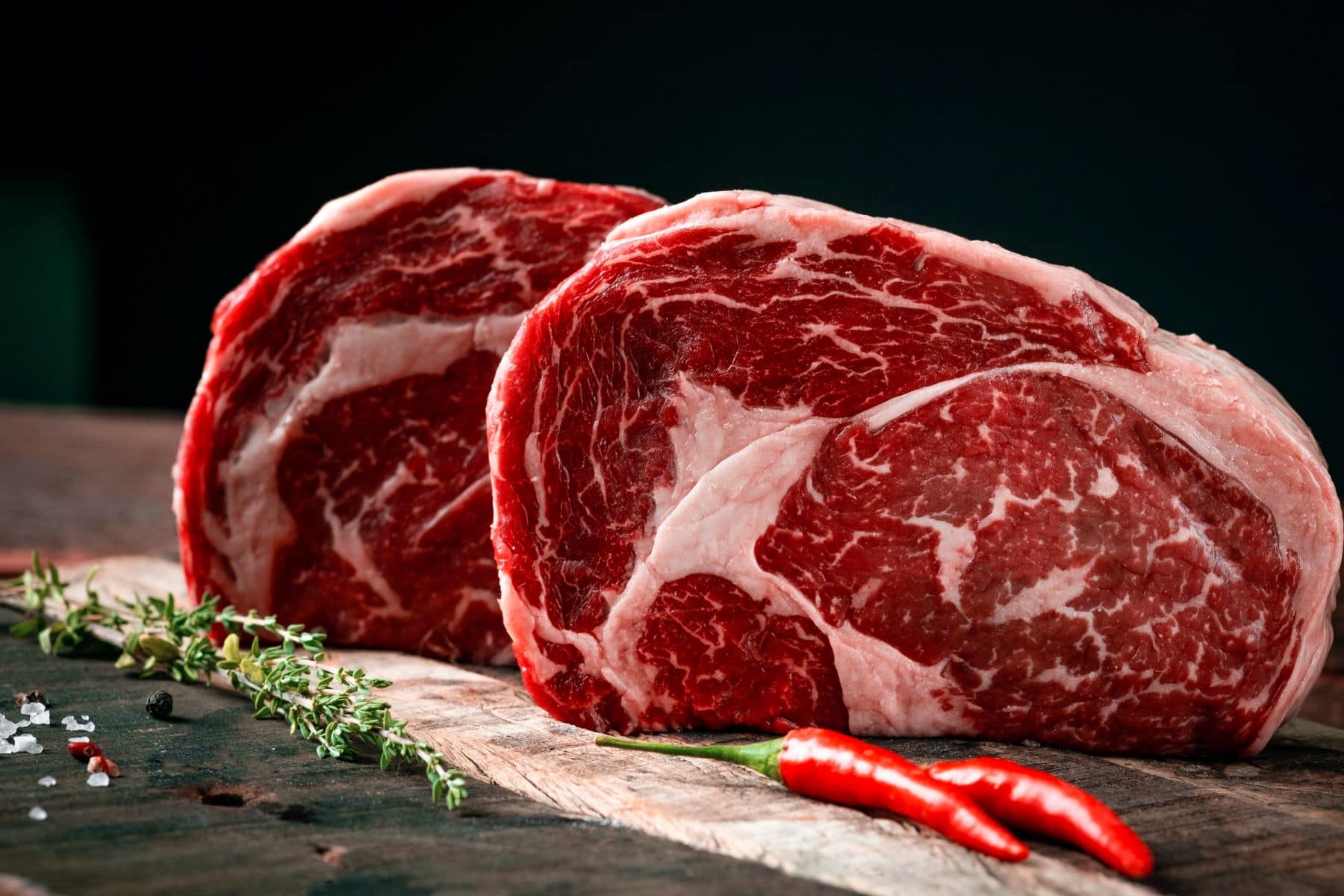 Zwei Rib Eye Steaks auf dunklem Holz. Im Bild Gewürze, Chilis und Kräuter. Frontalansicht.