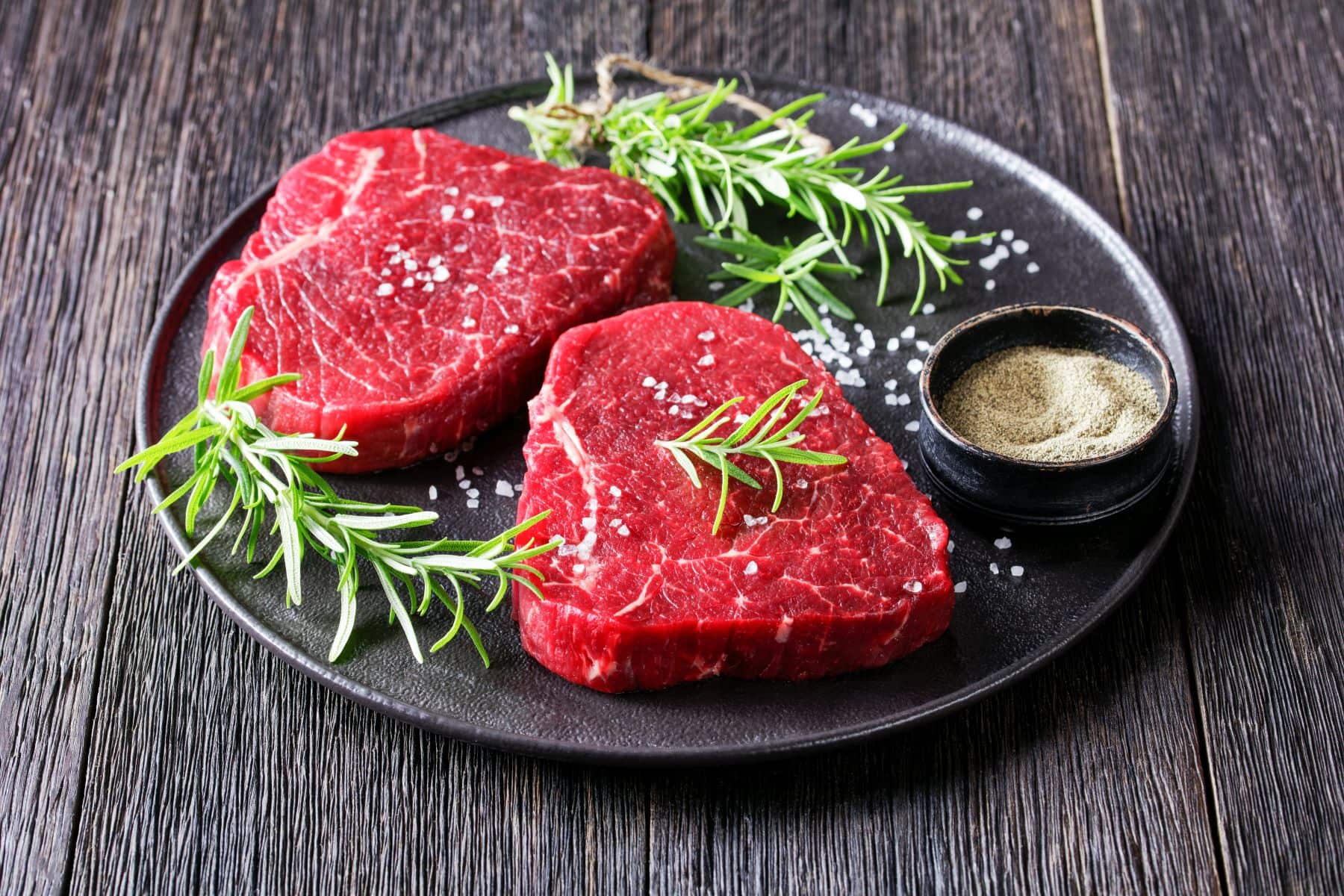 Zwei Rinderhüftsteak auf schwarzem Teller und dunklem Holz. Im Bild eine Schale mit Salz und Rosmarinzweige. Draufsicht.