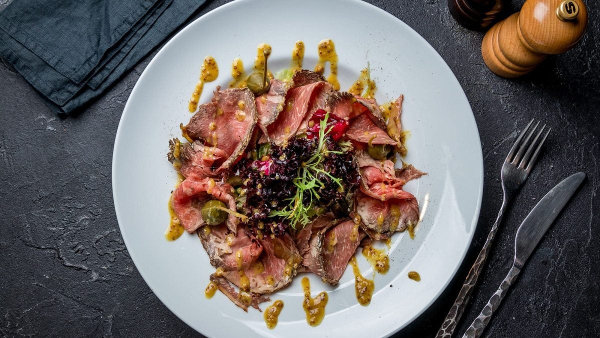 Draufsicht. Auf dunklem Untergrund steht ein weißer Teller mit einem Roastbeef-Salat mit Senf-Dressing. Daneben liegt Besteck.