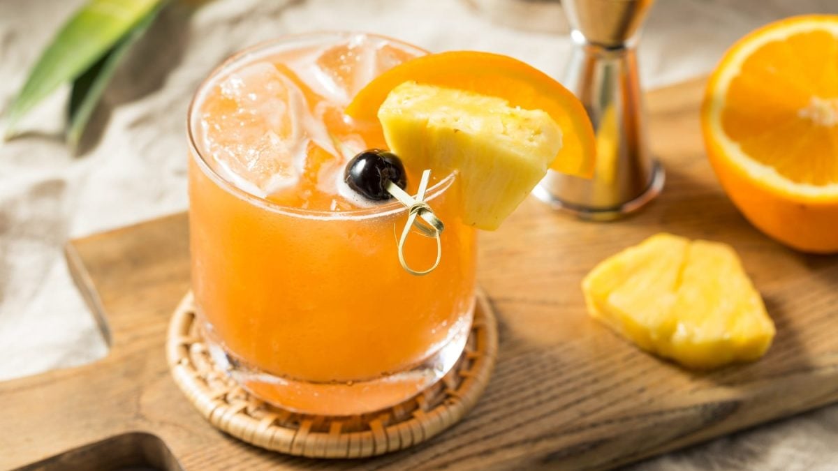 Ein orangefarbener Cocktail auf einem Holzbrett mit Ananas und Orange. Alles auf beigem Untergrund seitlich fotografiert.