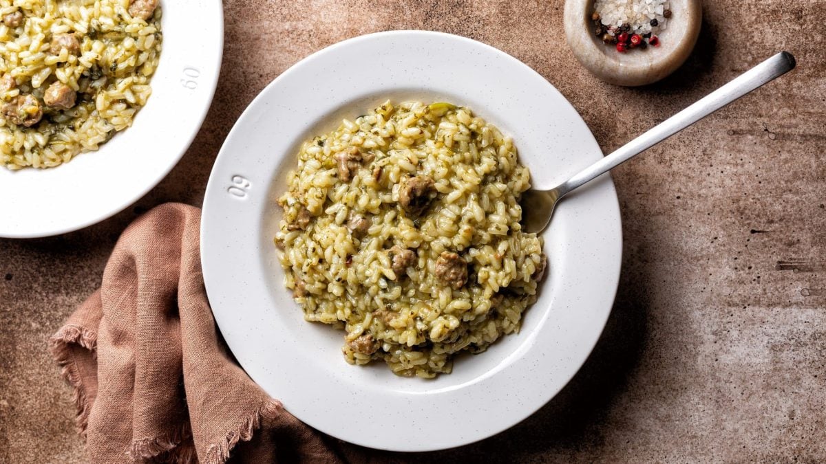 Draufsicht: Untergrund aus Holz mit zwei Tellern Salsiccia-Risotto. Daneben liegt ein Tuch und es steht eine Schale bunter Pfeffer auf dem Tisch.