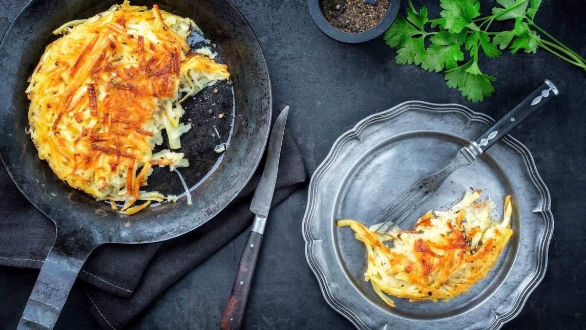 Draufsicht: Dunkler Untergrund, eine Gusseiserne Pfanne und eine Teller aus Metall mit einem Schweizer Rösti aus rohen Kartoffeln.