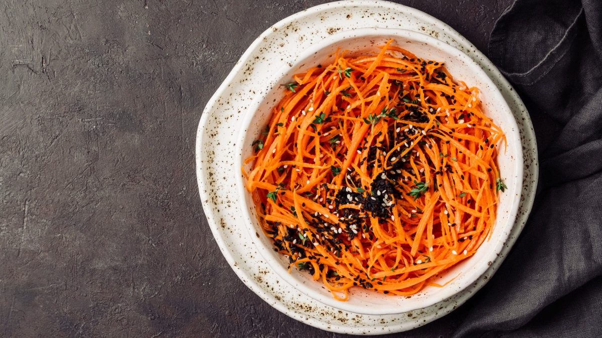 Draufsicht: Dunkler Untergrund und ein weißer Teller mit einem Sesam-Karotten-Salat mit Mandelsauce.