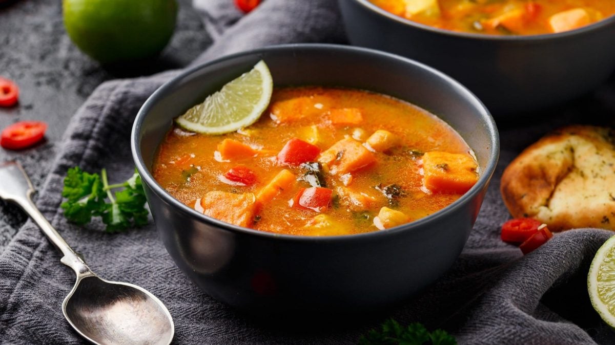 Eine schwarze Schale mit Süßkartoffel-Curry-Suppe, darin auch eine Limettenspalte, daneben ein Löffe, Chilistücke und geröstetes Brot.