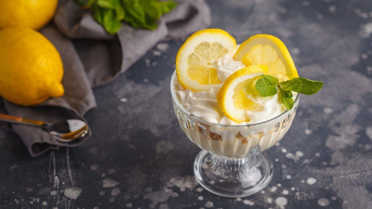 Ein Glas Syllabub, ein weiße Creme aus Zitrone, Weißwein und Sahne, garniert mit Minze und Zitronenscheiben, daneben liegen Zitronen.