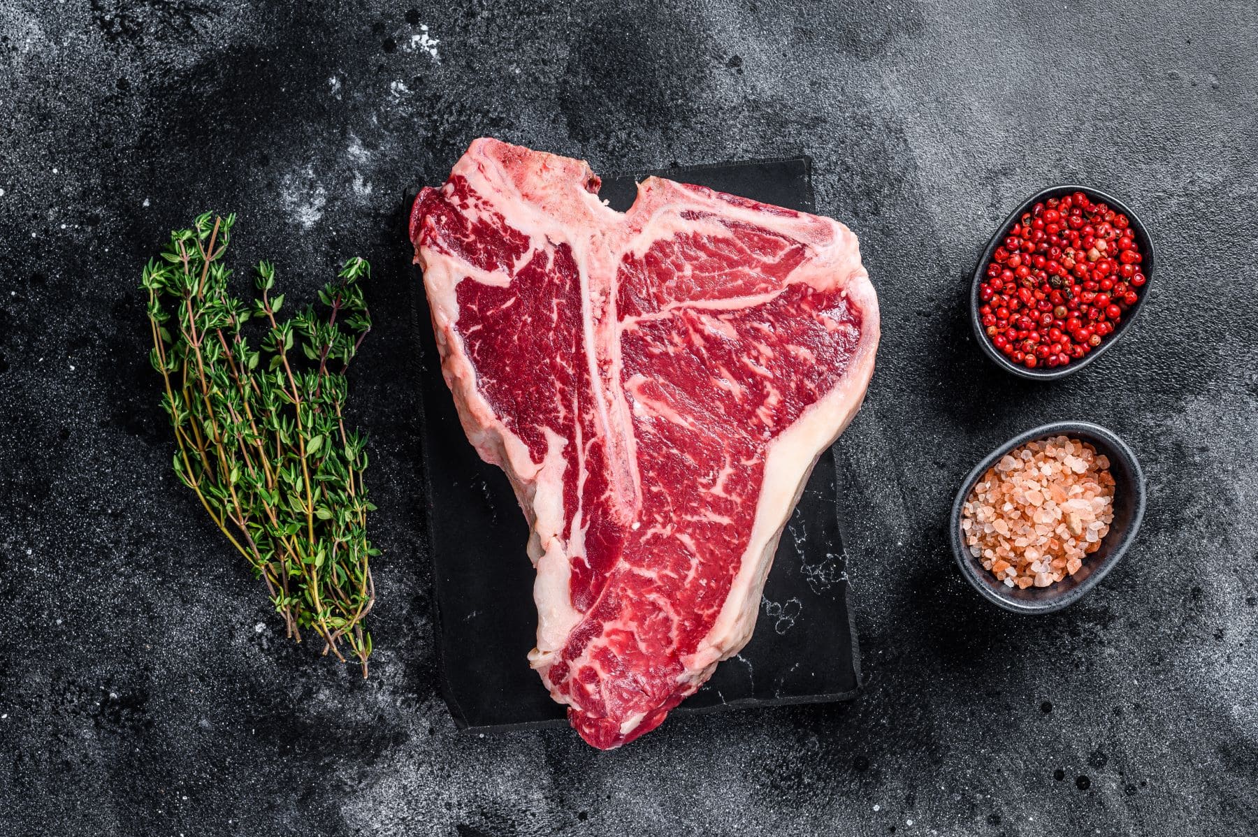 T-Bone-Steal auf dunklem Stein mit Thymianzweig und zwei Schalen mit Gewürzen. Draufsicht.