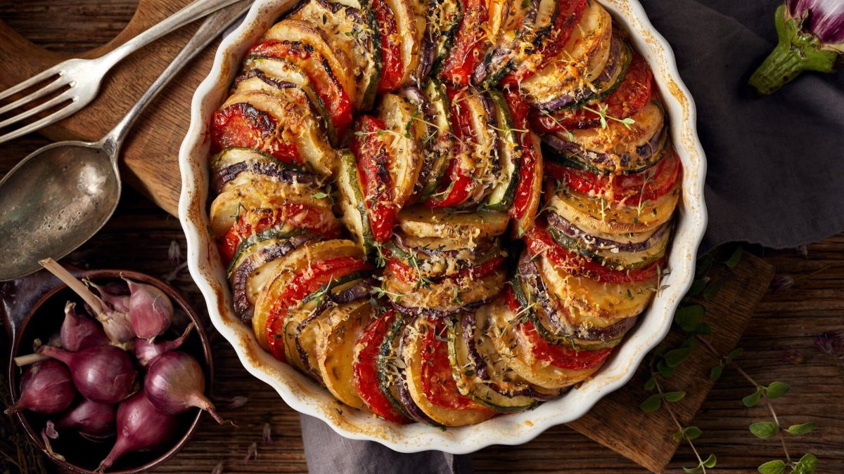 Eine runde Auflaufform Tian, französischer Gemüseauflauf, bei dem Tomaten, Zucchini und Aubergine fächerartig angeordnet werden. Hier in der Draufsicht und mit Käse überbacken, daneben eine Schale mit Schalotten.