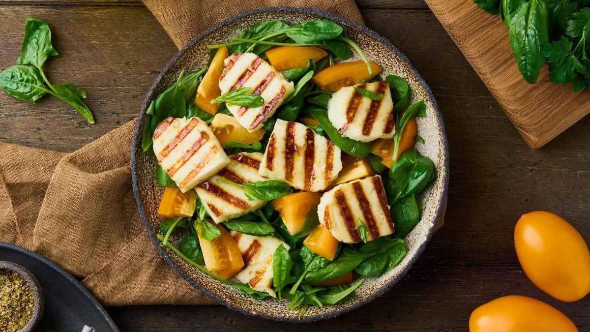 Draufsicht: eine gegrillter Tomaten-Halloumi-Salat auf Holzuntergrund. Daneben liegen gelbe Tomaten und Spinatblätter.