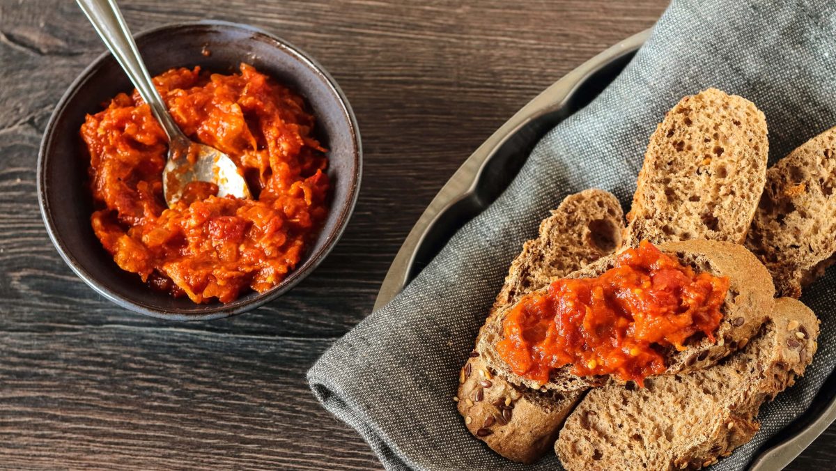 Draufsicht: Dunkler Untergrund, eine dunkle, kleine Schale mit einem veganen Tofu-Tomaten-Aufstrich. Auf einem grauen Tuch liegen mehrere Scheiben Brot.
