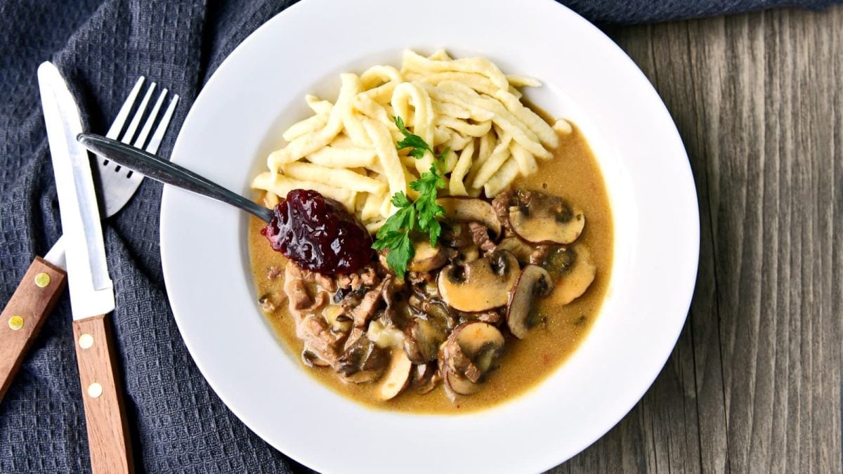 Ein veganes Champignon-Rahm-Geschnetzeltes mit Spätzle in einem weißen Teller vor einem hölzernen Hintergrund. Links davon ein schwarzes Geschirrtuch, darauf ein Messer und eine Gabel.