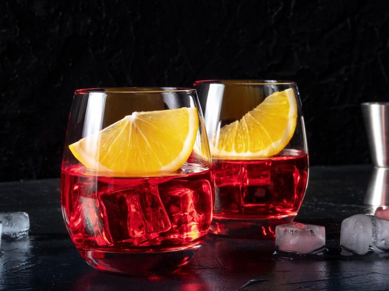 Zwei Gläser Wermut-Spritz mit Eiswürfeln und je einer Orangenspalte vor dunklem Hintergrund, daneben ein Cocktail-Messbecher.