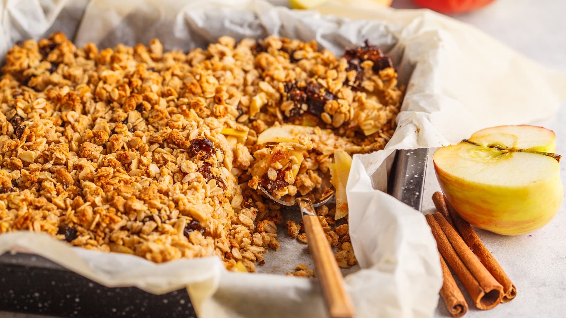 Ein Blech mit dem Apfel-Crumble-Cake auf einem hellen tisch mit Zimtstangen. Alles seitlich und nah fotografiert.