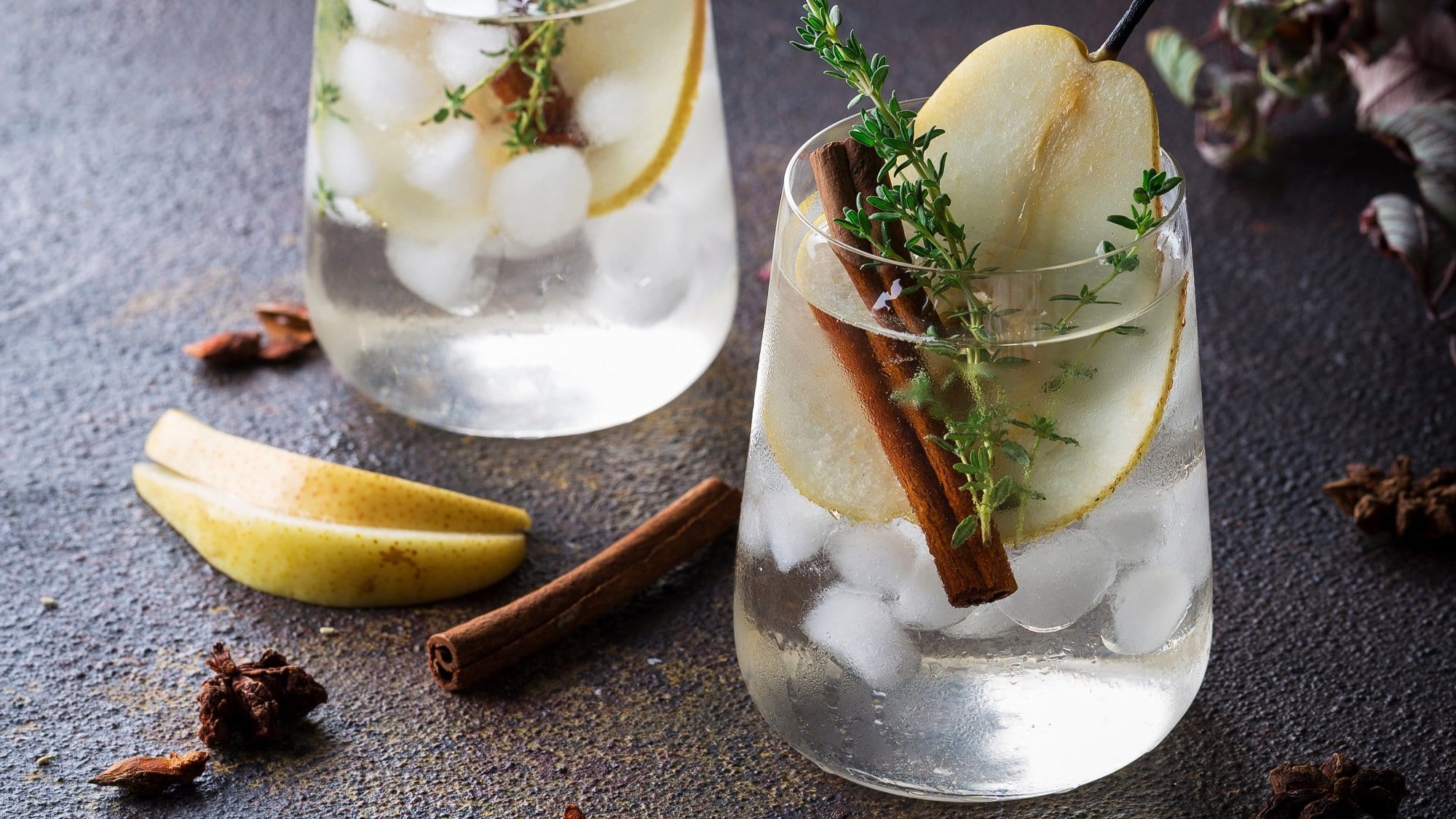 2 Gläser Autumn Pear Cocktail mit Eiswürfeln, einer BIrnenscheibe, einer Zimtstange und Thymian garniert, drumherum Zimtstangen und BIrnenscheiben.