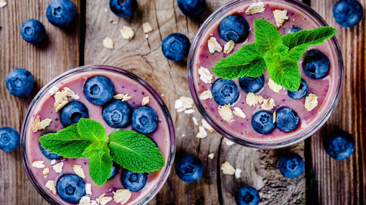 Draufsicht: Untergrund aus Holz, zwei Gläser Blaubeeren-Bananen-Smoothie mit Haferflocken und Minze. Drum herum liegen Blaubeeren.