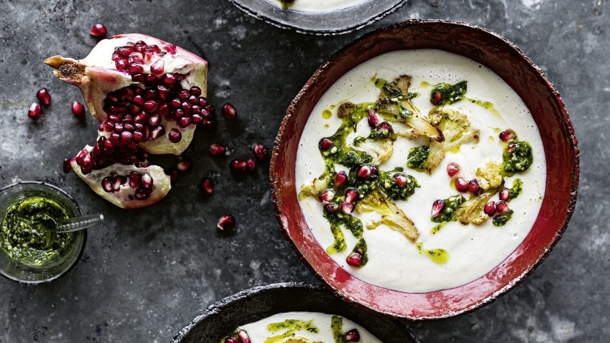 Eine Schüssel Blumenkohlsuppe mit Tahin und Granatapfelpesto, daneben ein geöffneter Granatapfel und zwei weitere Schüsseln, die man nur angeschnitten sieht, Draufsicht.
