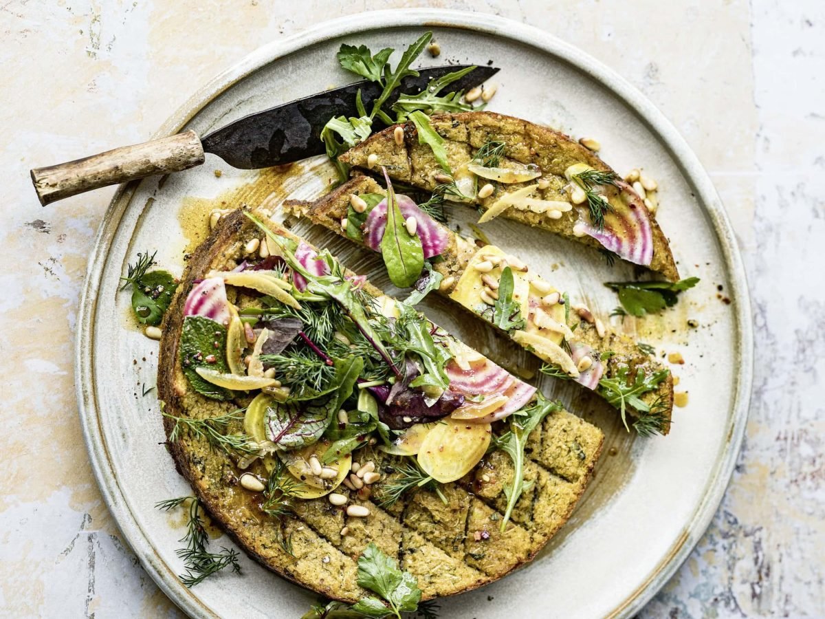 Ein angeschnittener Bulgurkuchen mit Kräutersalat auf einem Teller, auf dem ein Messer liegt. Das Rezept stammt aus dem Kochbuch "Deftig vegan Orient"