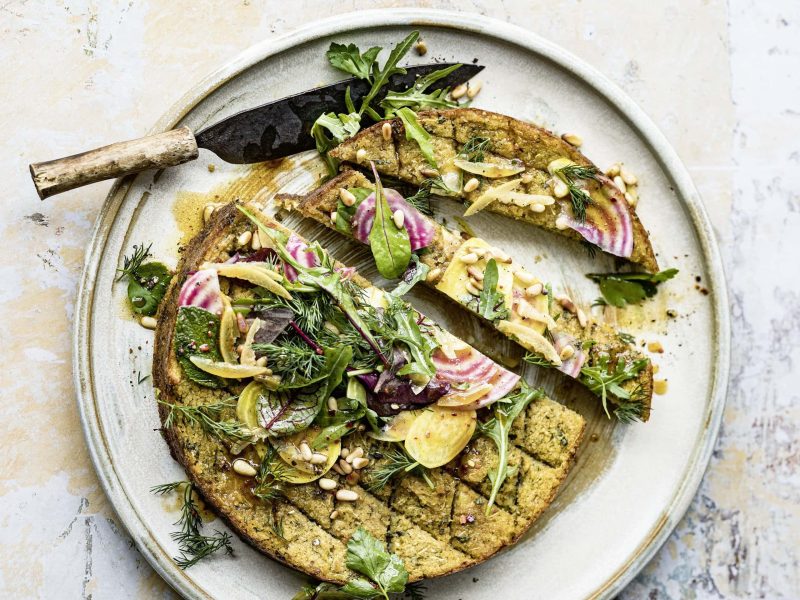 Ein angeschnittener Bulgurkuchen mit Kräutersalat auf einem Teller, auf dem ein Messer liegt. Das Rezept stammt aus dem Kochbuch "Deftig vegan Orient"