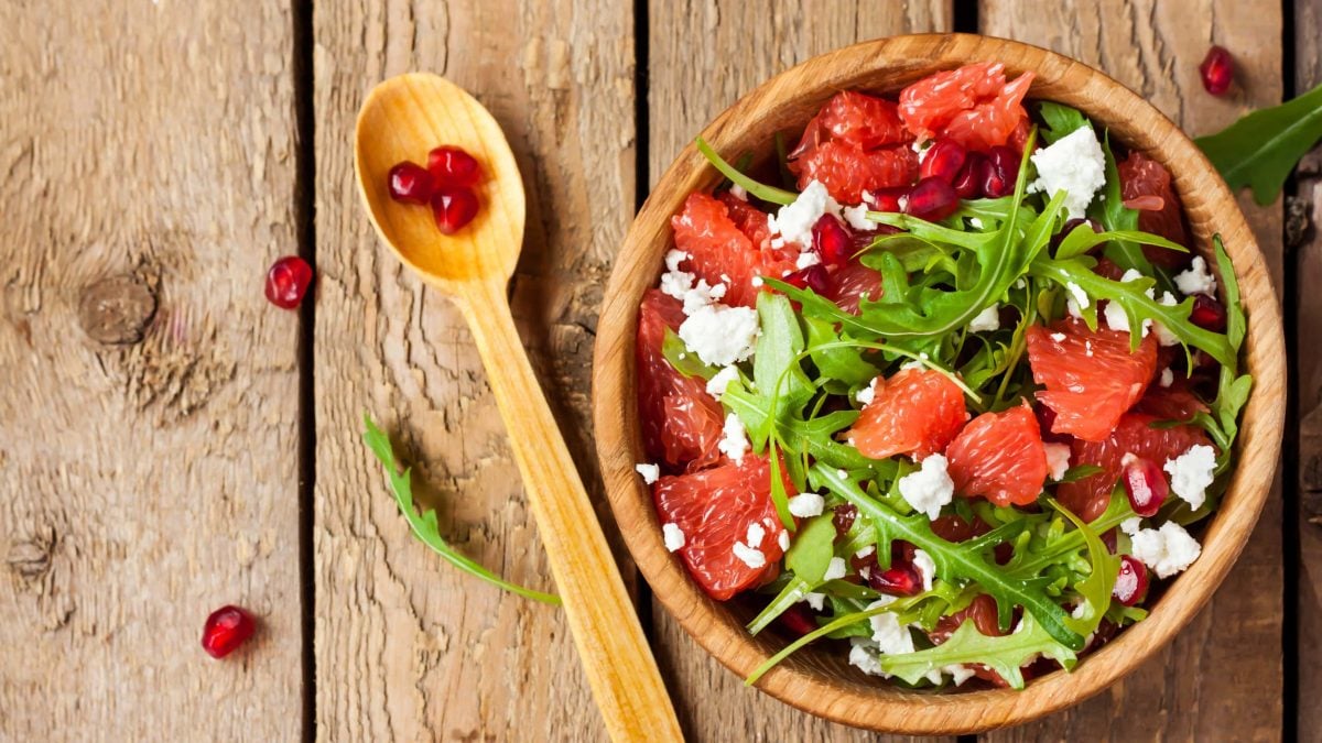 Draufsicht: Eine Schüssel Grapefruit-Salat mit Feta und Granatapfelkernen auf einem Untergrund aus Holz. Daneben liegt ein Löffel aus Holz.
