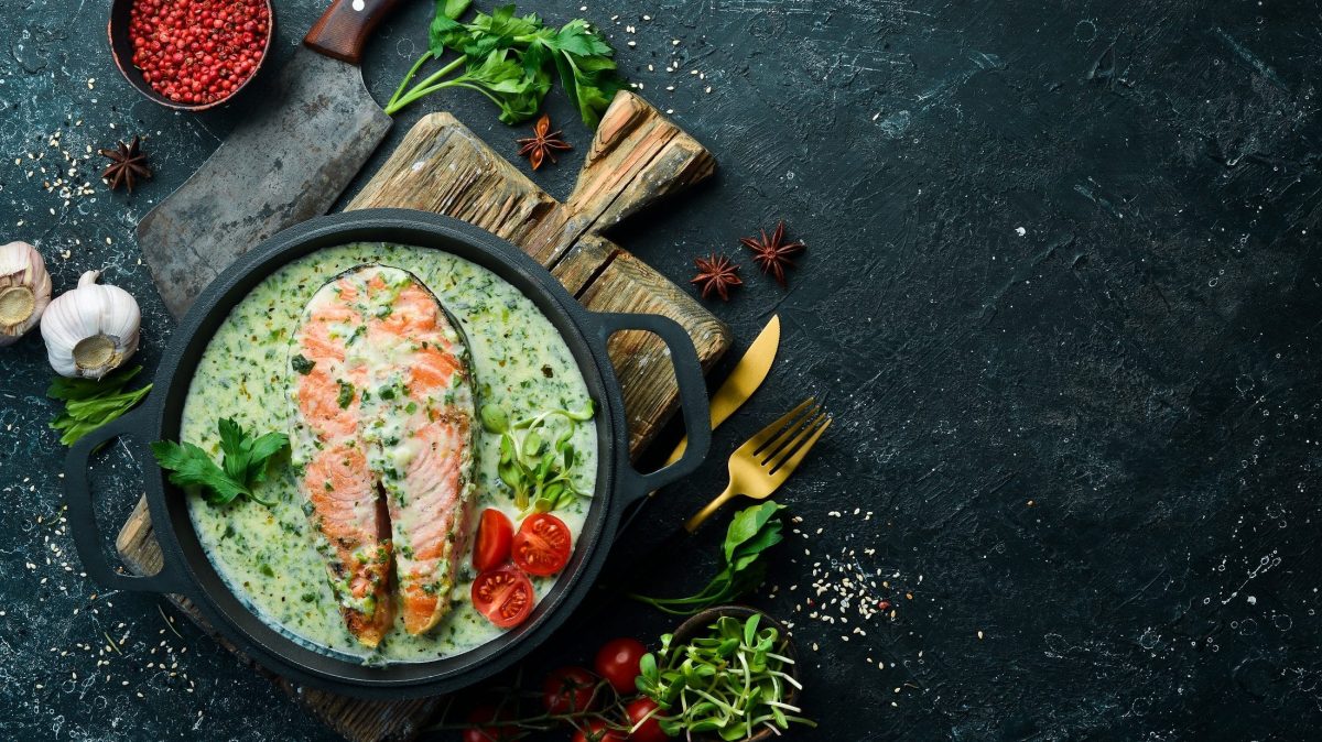Draufsicht: Auf dunklem Untergrund steht eine Pfanne mit Lachs in Spinat-Sahnesauce. Drum herum liegen Kräuter.