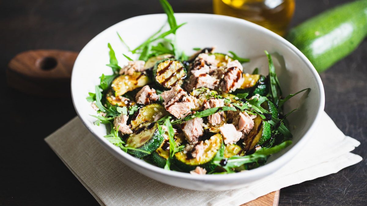 Draufsicht: eine weiße Schüssel Thunfisch-Salat mit gegrillter Zucchini auf einem Tuch, das auf einem Holzbrettchen liegt. Im Hintergrund ein Teil einer Zucchini und eine Flasche Öl.