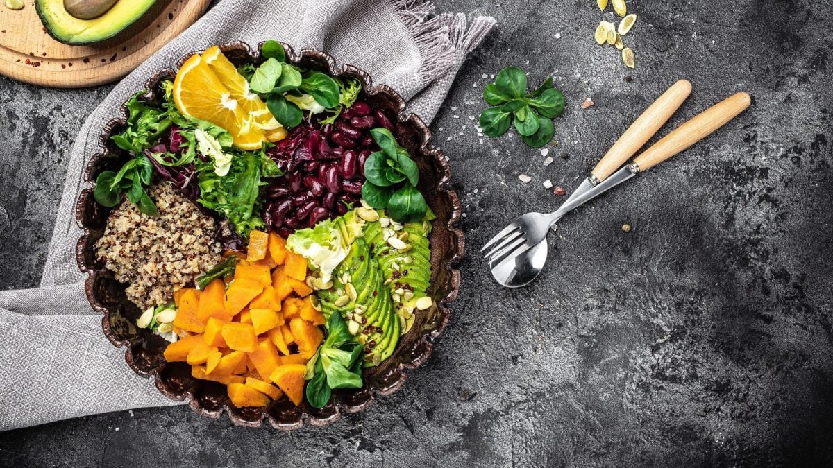Fitness-Bowl mit Avocado, Feldsalat, Quinoa, Süßkartoffel, roten Bohnen und Orangenscheiben in der Draufsicht, daneben Besteck und ein paar Zutaten.