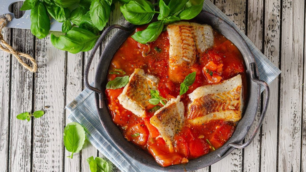 Draufsicht: auf einem hellen Holztisch steht eine Pfanne geschmortes Fischfilet auf Tomatensauce. Daneben liegen viele frische Basilikumblätter.