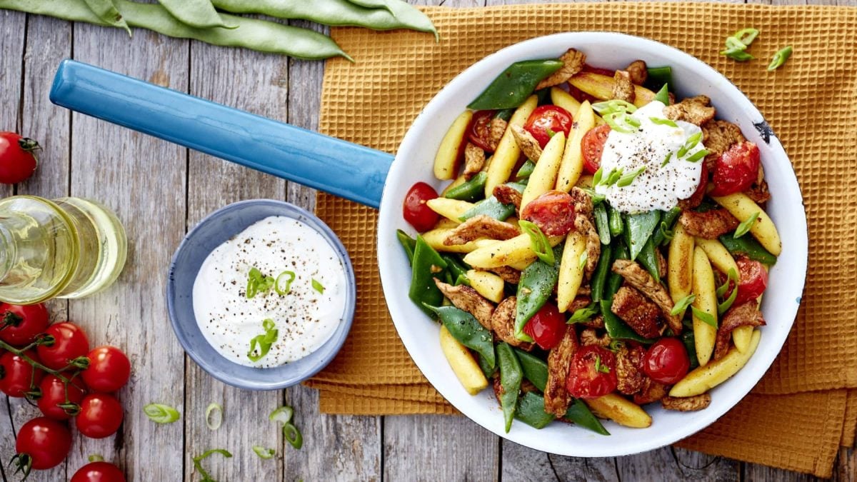 Eine Gyros-Schupfnudel-Pfanne auf einem gelben Geschirrtuch auf hölzernem Hintergrund. Links ein Schüsselchen mit Sour Cream, Tomaten und Öl. Oben auf dem Bild Bohnen.