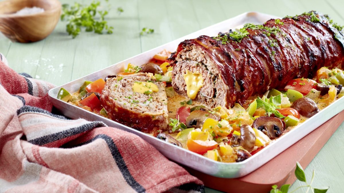 Ein gefüllter Hackbraten in Paprika-Rahm auf einem Blech auf einem grünen Untergrund. Links ein rotes Geschirrtuch, links oben Salz und frische Kräuter.