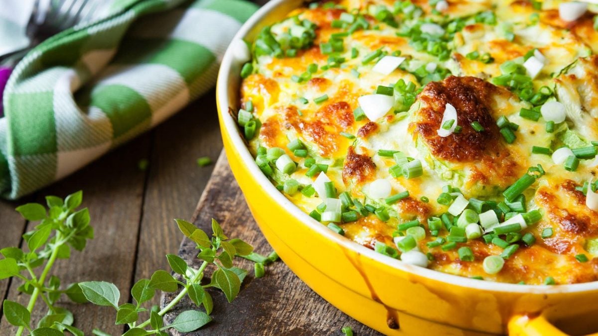 Herbstlicher Gemüseauflauf mit Käse überbacken und Frühlingszwiebeln bestreut, daneben Kräuter und ein grün-weiß kariertes Geschirrtuch.