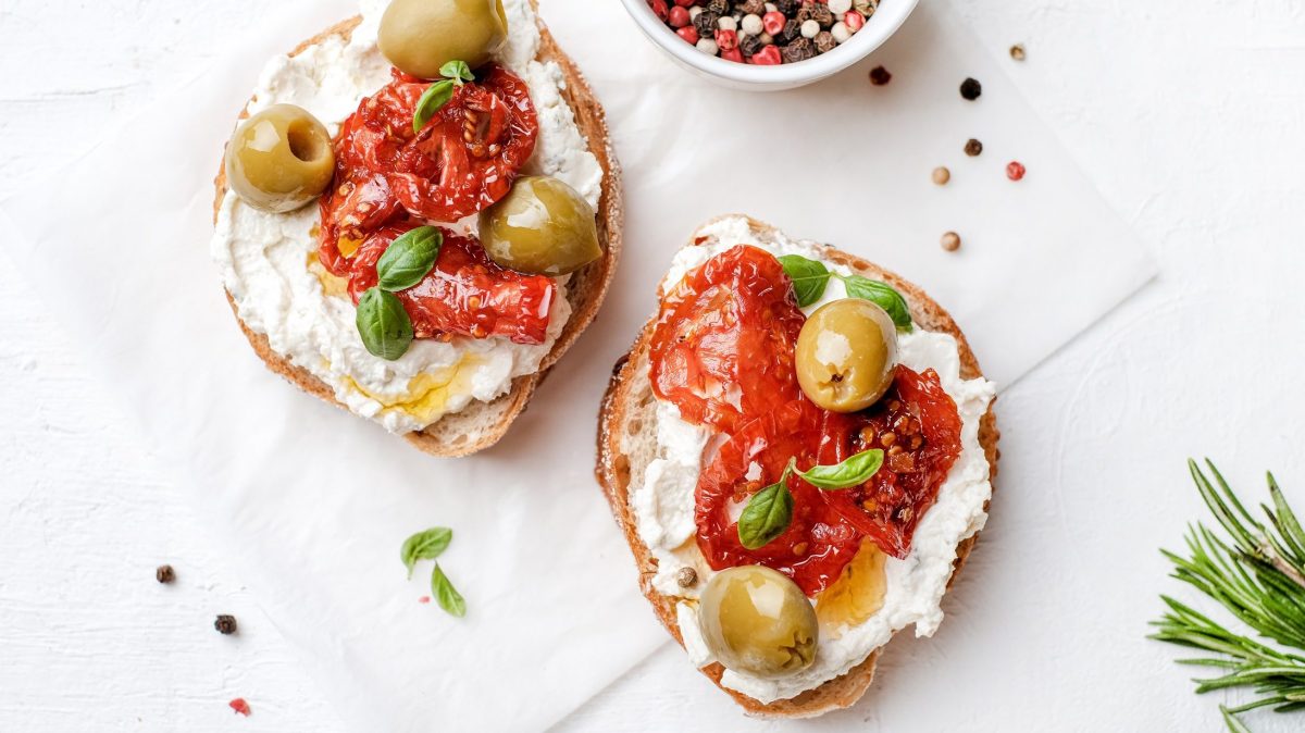 Draufsicht: Weißer Untergrund und zwei Käse-Bruschetta mit getrockneten Tomaten und Oliven.