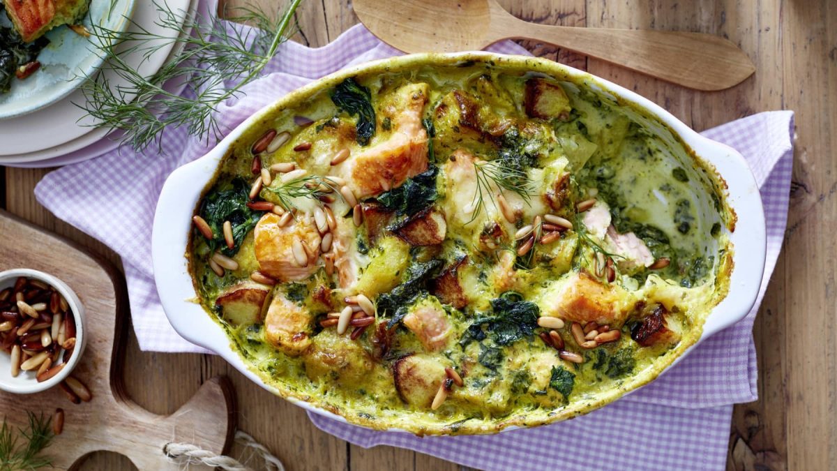 Ein Kartoffel-Lachs-Auflauf mit Spinat in einer weißen Auflaufform auf einem lila-kartierten Geschirrtuch auf einem hölzernen Hintergrund. Links ein Schneidebrett, Pinienkerne, Teller und Dill. Oben auf dem Bild ein Holzlöffel.