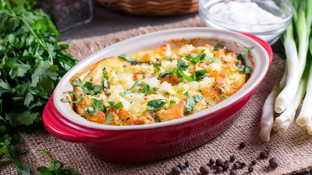 Ein Kürbis-Gnocchi-Auflauf in einer ovalen, roten Auflaufform, daneben Zutaten wie Frischkäse in einer Glasschale, Petersilie oder Lauchzwiebeln.