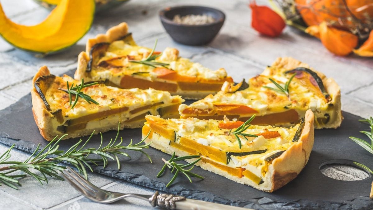 4 Stücke Kürbis-Quiche auf einer Schieferplatte, mit Rosmarin belegt, drumherum Kürbisstücke und Kräuter.